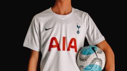 Pemain sepak bola dewasa dengan jersey Tottenham Hotspur memegang bola di latar belakang gelap.
