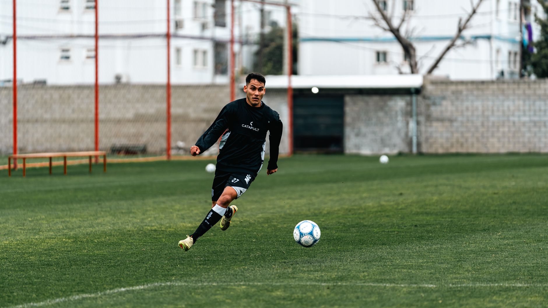 Gambar dinamis dari pemain sepak bola yang mengenakan pakaian olahraga menggiring bola di lapangan rumput saat latihan di siang hari.