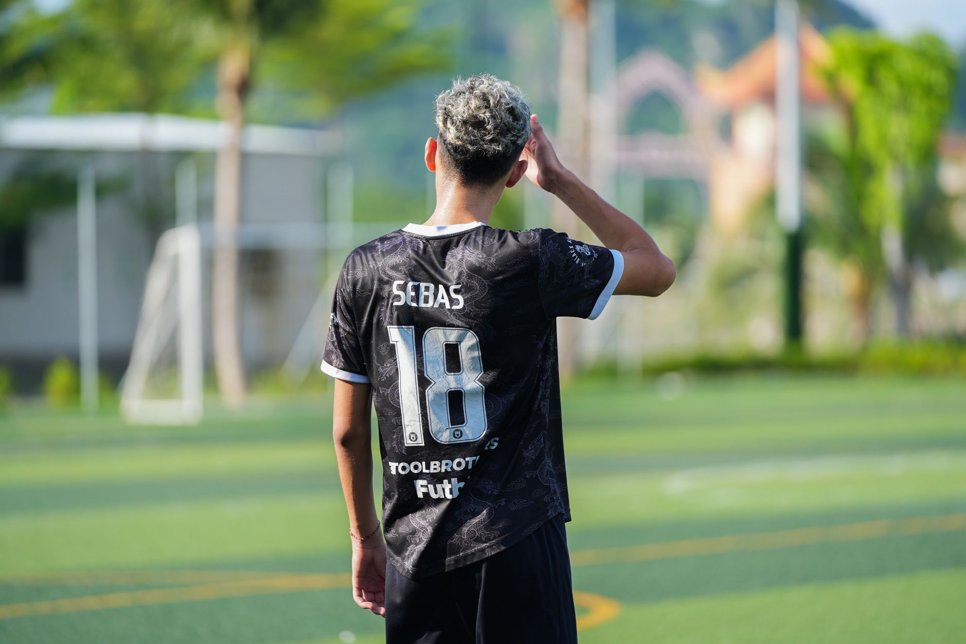 Pemain muda dengan jersey hitam di lapangan sepak bola luar ruangan pada siang hari, menghadap menjauh dari kamera.