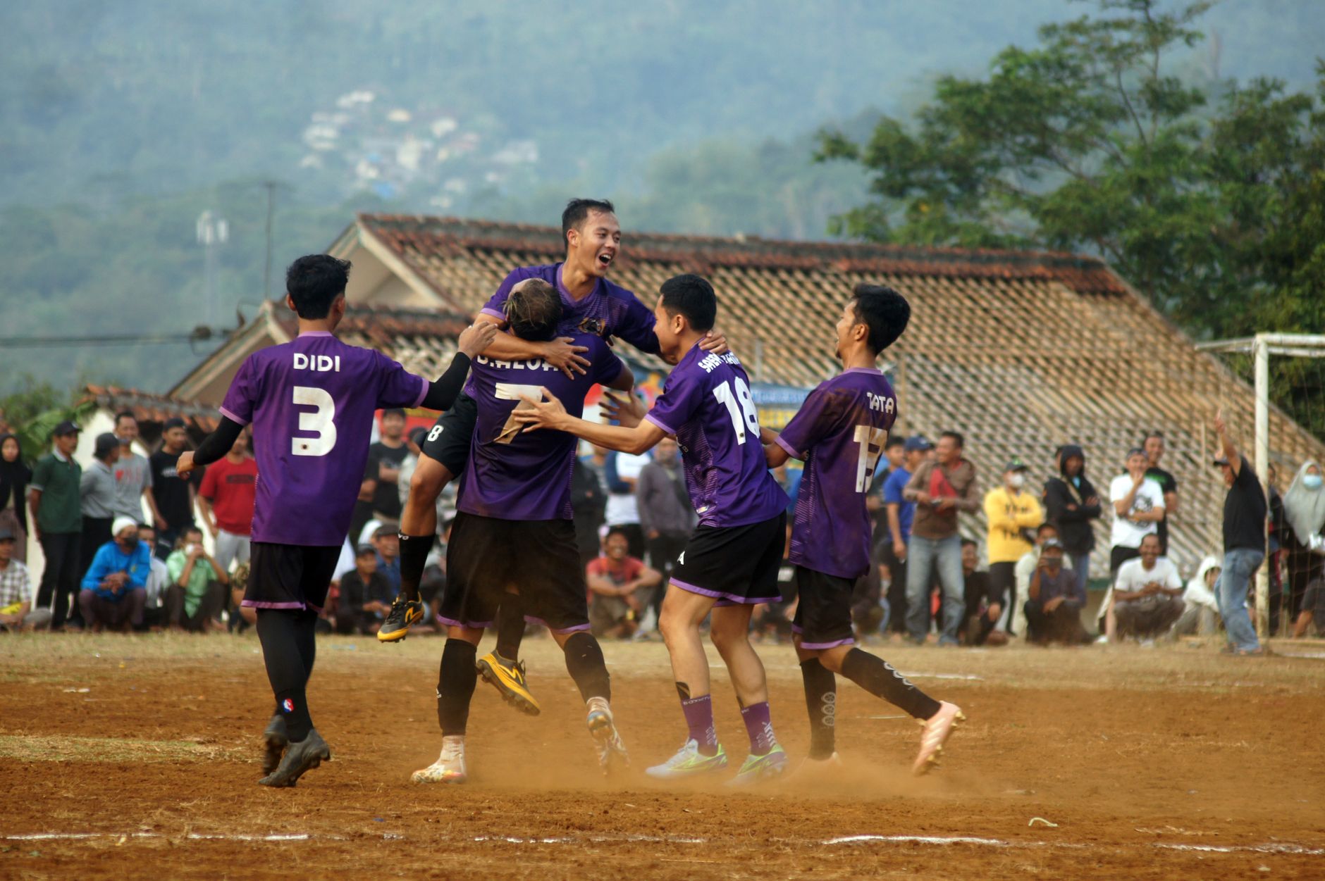 Pemain sepak bola merayakan gol dalam pertandingan luar ruangan yang berdebu dengan penonton.