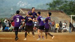 Pemain sepak bola merayakan gol dalam pertandingan luar ruangan yang berdebu dengan penonton.