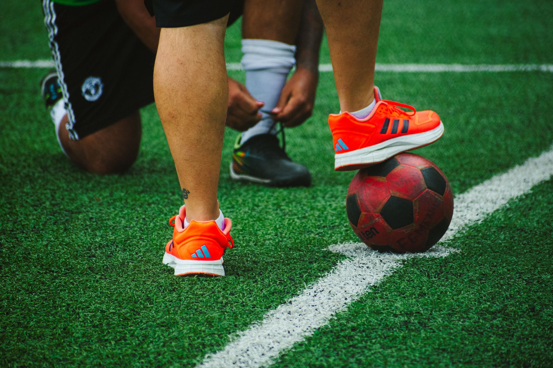 Close-up pemain sepak bola saat latihan, satu mengikat tali sepatu, yang lain menyeimbangkan bola di rumput.