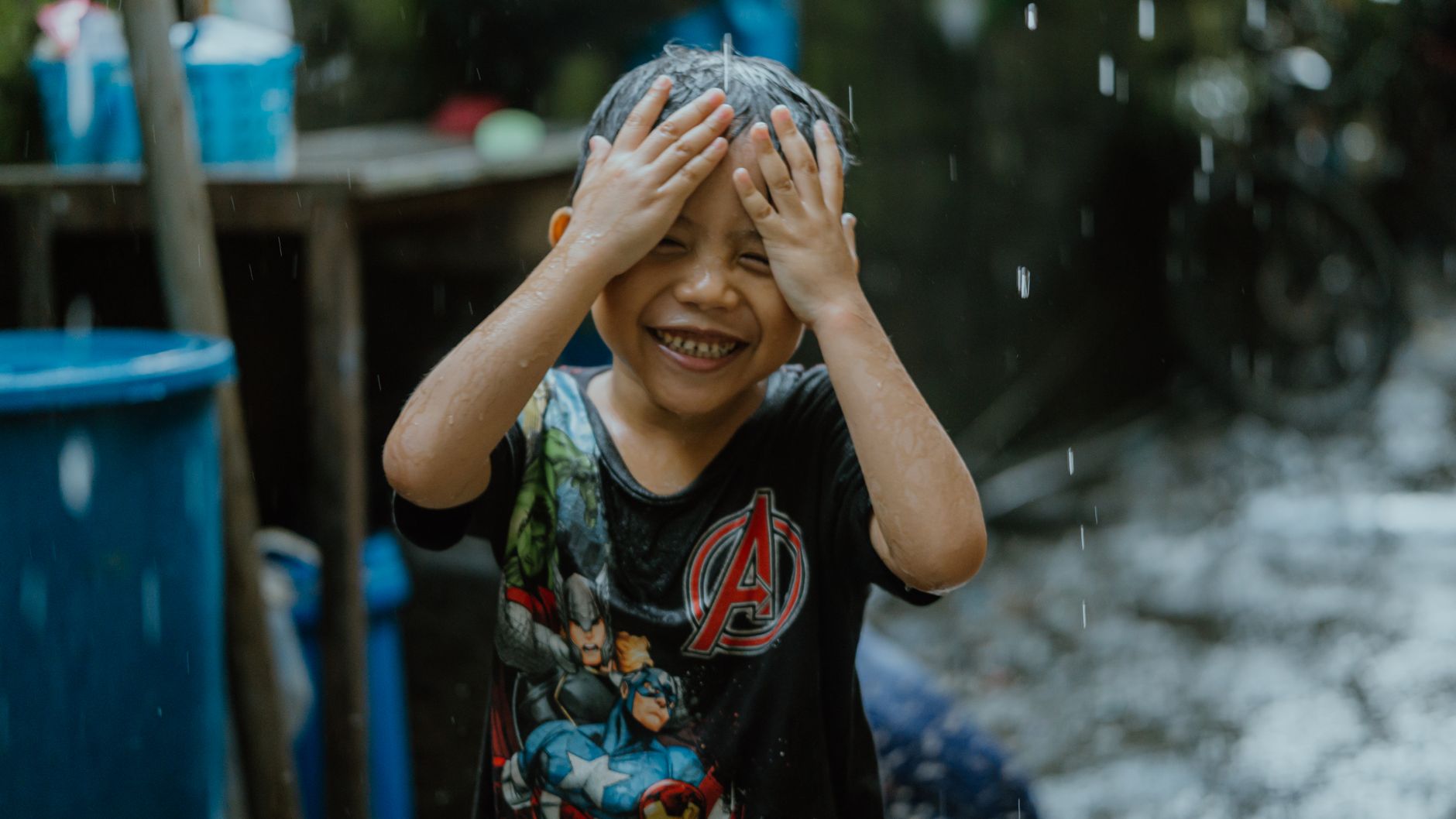 Anak yang tersenyum menikmati hujan dengan pakaian santai, merangkul momen masa kecil yang bebas.
