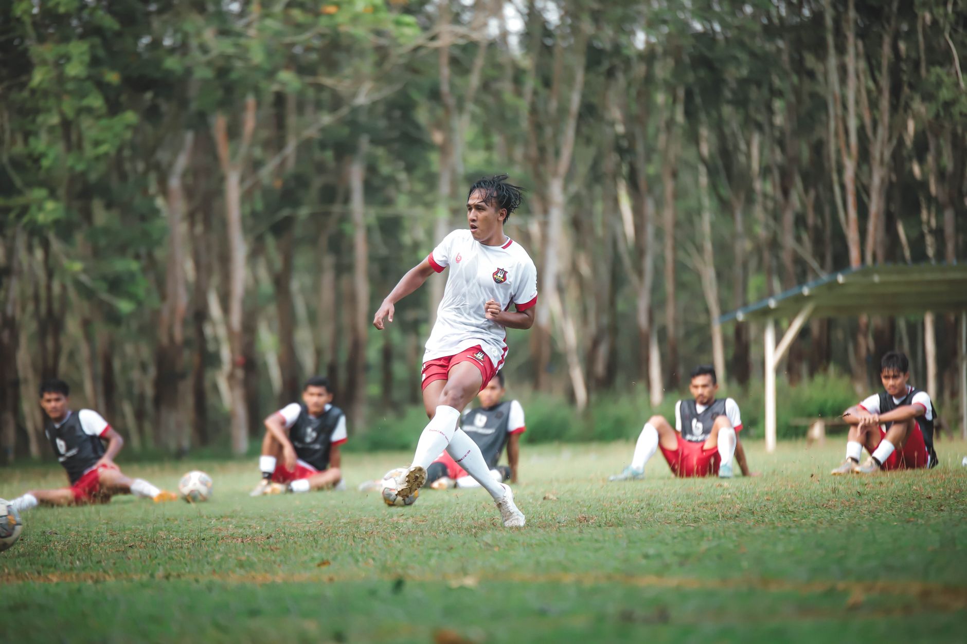 Pemain sepak bola yang dinamis dalam aksi selama sesi pelatihan luar ruangan dengan rekan tim di latar belakang.