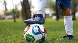 Seorang pemain sepak bola muda yang mengenakan kaus kaki putih berdiri di atas bola sepak warna-warni di luar ruangan.