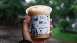 Close-up tangan memegang cangkir kopi es bermerek di luar ruangan, dengan latar belakang hijau buram yang cerah.