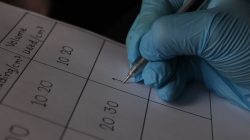 Gambar close-up seorang ilmuwan yang mengenakan sarung tangan biru sedang menuliskan pengukuran pada lembar data.