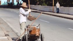Seseorang sedang menyapu jalan dengan sapu dan kereta di lingkungan perkotaan.