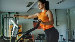 Seorang wanita yang fokus berolahraga menggunakan alat elliptical trainer di pusat kebugaran, melambangkan kebugaran dan tekad.