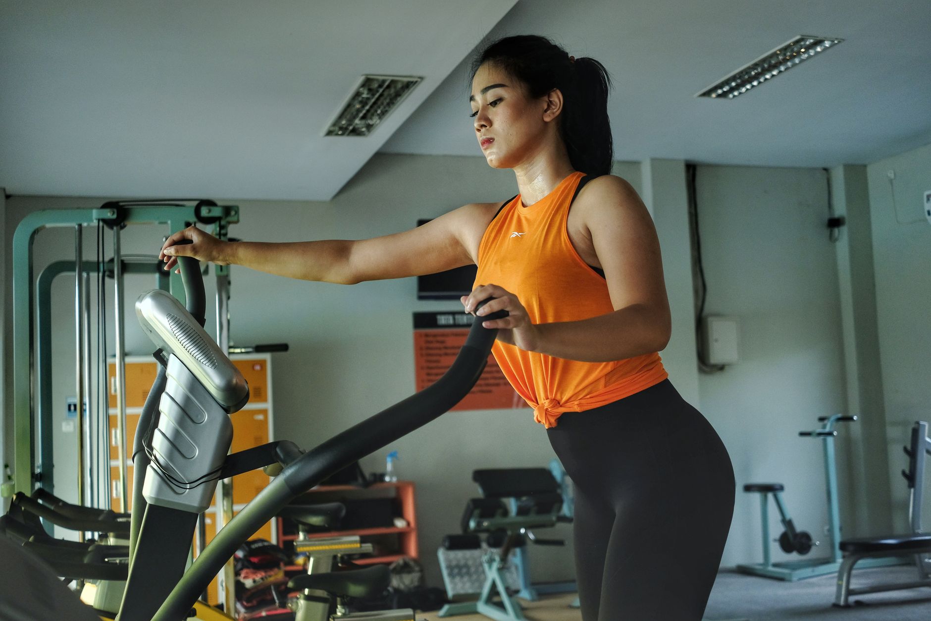 Seorang wanita yang fokus berolahraga menggunakan alat elliptical trainer di pusat kebugaran, melambangkan kebugaran dan tekad.