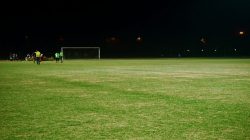 Pemandangan lapangan sepak bola di malam hari dengan pemain dan tiang gawang di bawah lampu sorot.