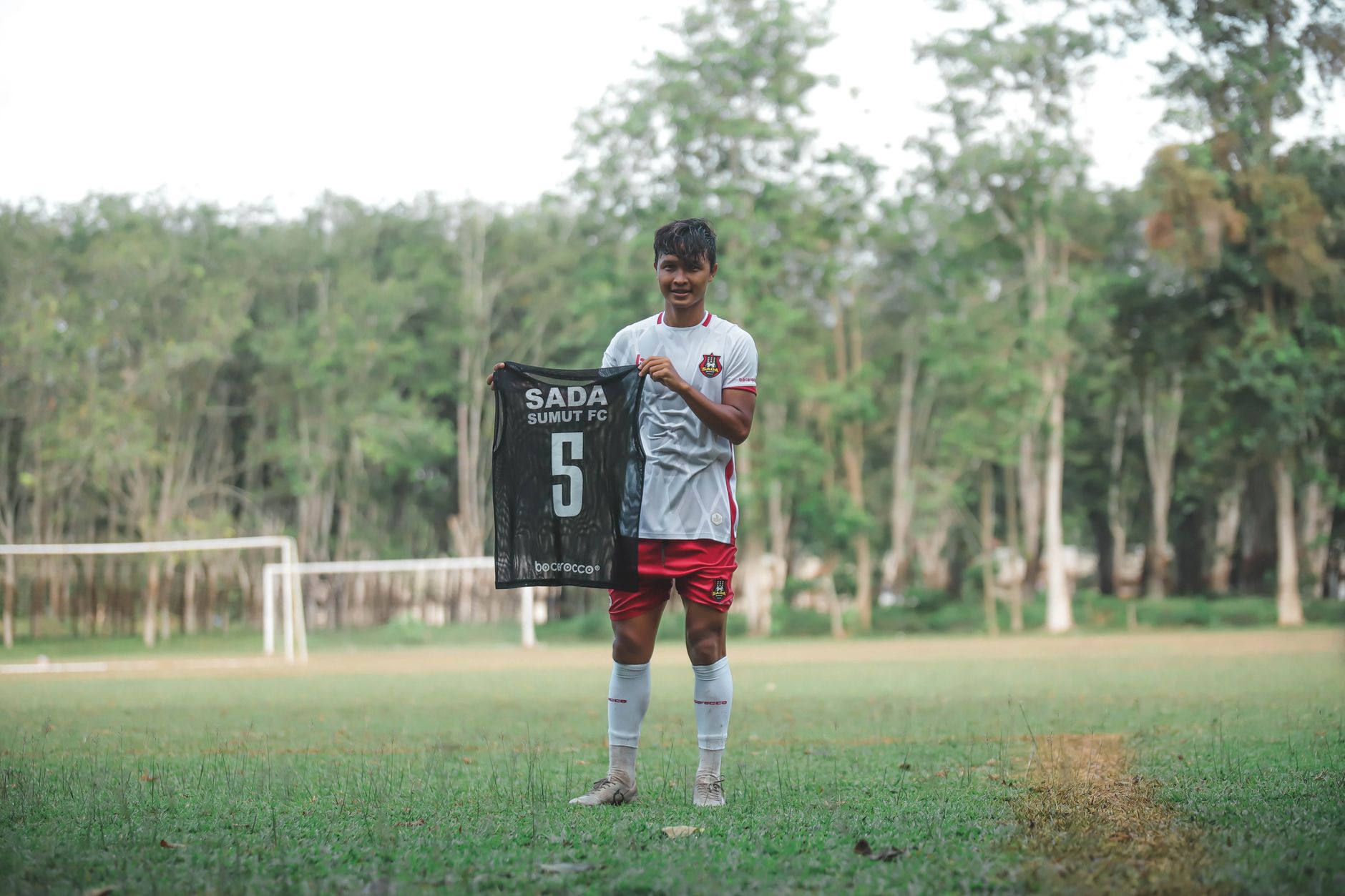 Seorang pemain sepak bola muda dengan bangga memegang jersey tim di lapangan hijau yang dikelilingi pohon.