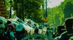 Sebuah adegan dinamis orang-orang dan polisi selama protes jalanan di area perkotaan yang cerah.