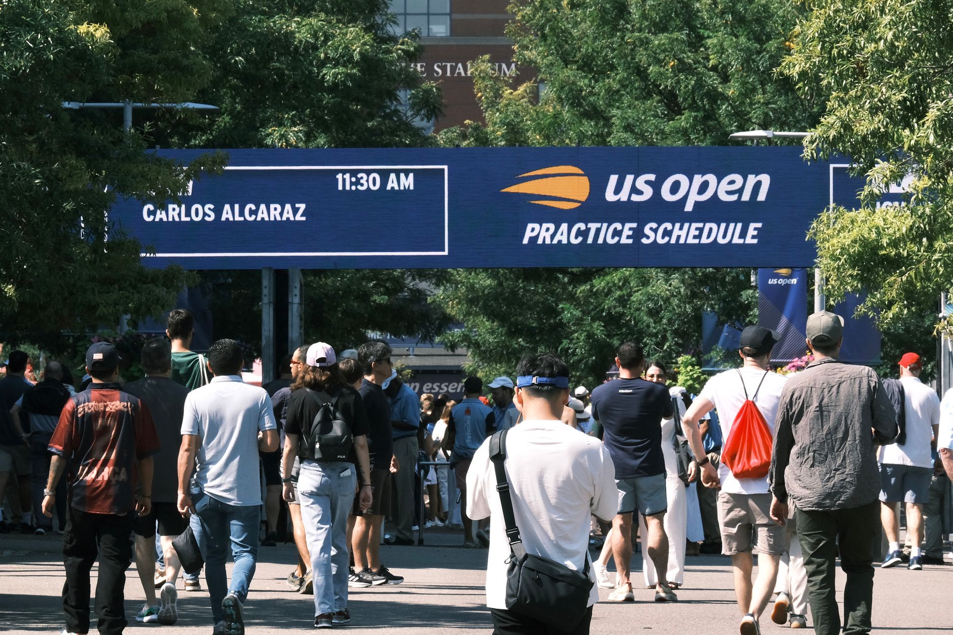 Orang-orang berkumpul di papan jadwal latihan US Open di bawah langit yang cerah.