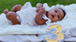 Bayi lucu berbaju motif bunga berbaring di atas selimut di luar ruangan di samping angka tiga yang dekoratif.