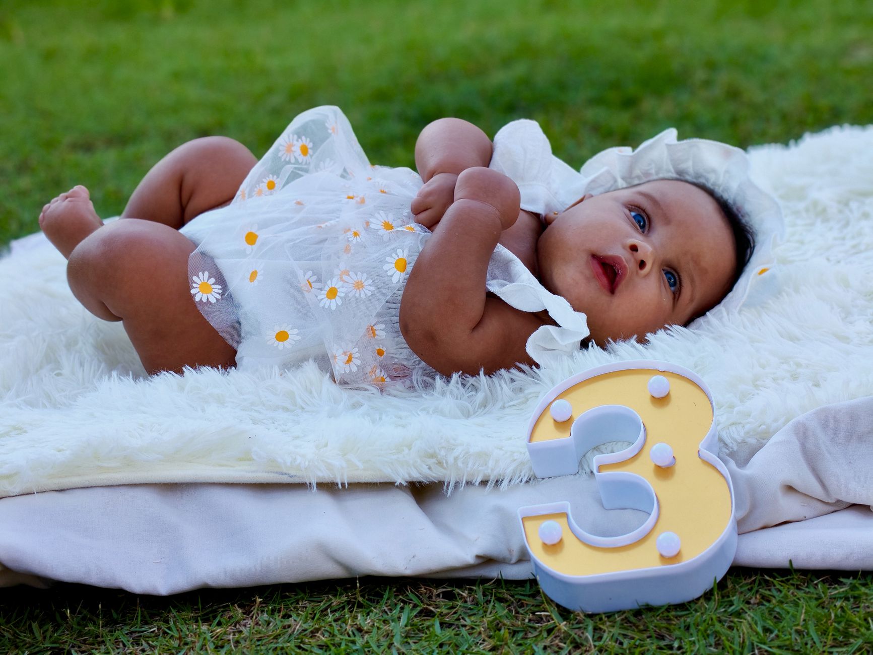 Bayi lucu berbaju motif bunga berbaring di atas selimut di luar ruangan di samping angka tiga yang dekoratif.